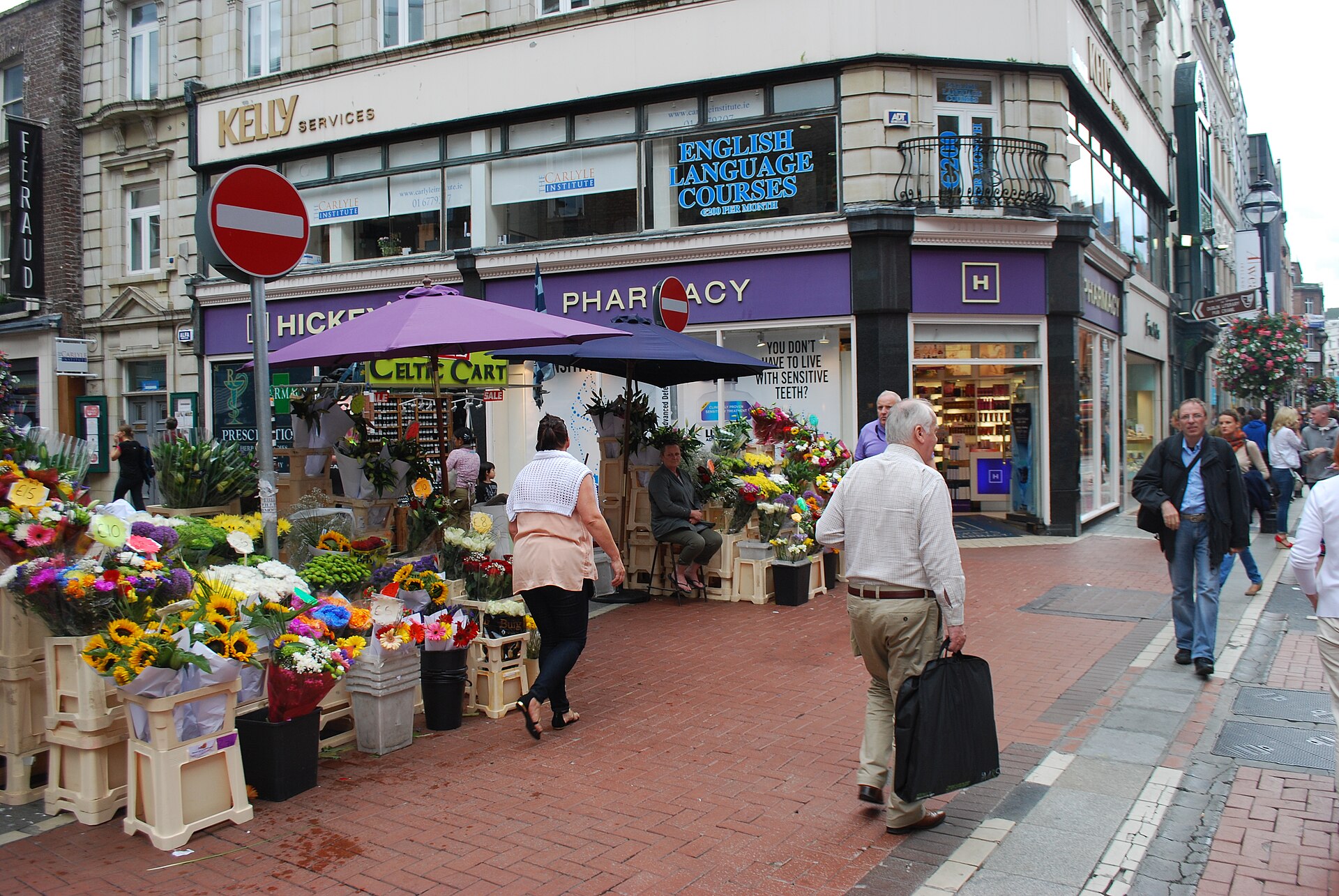 Pedestrians pass buckets of colourful flowers beneath a purple parasol at a shopfront corner on Grafton Street, Dublin.