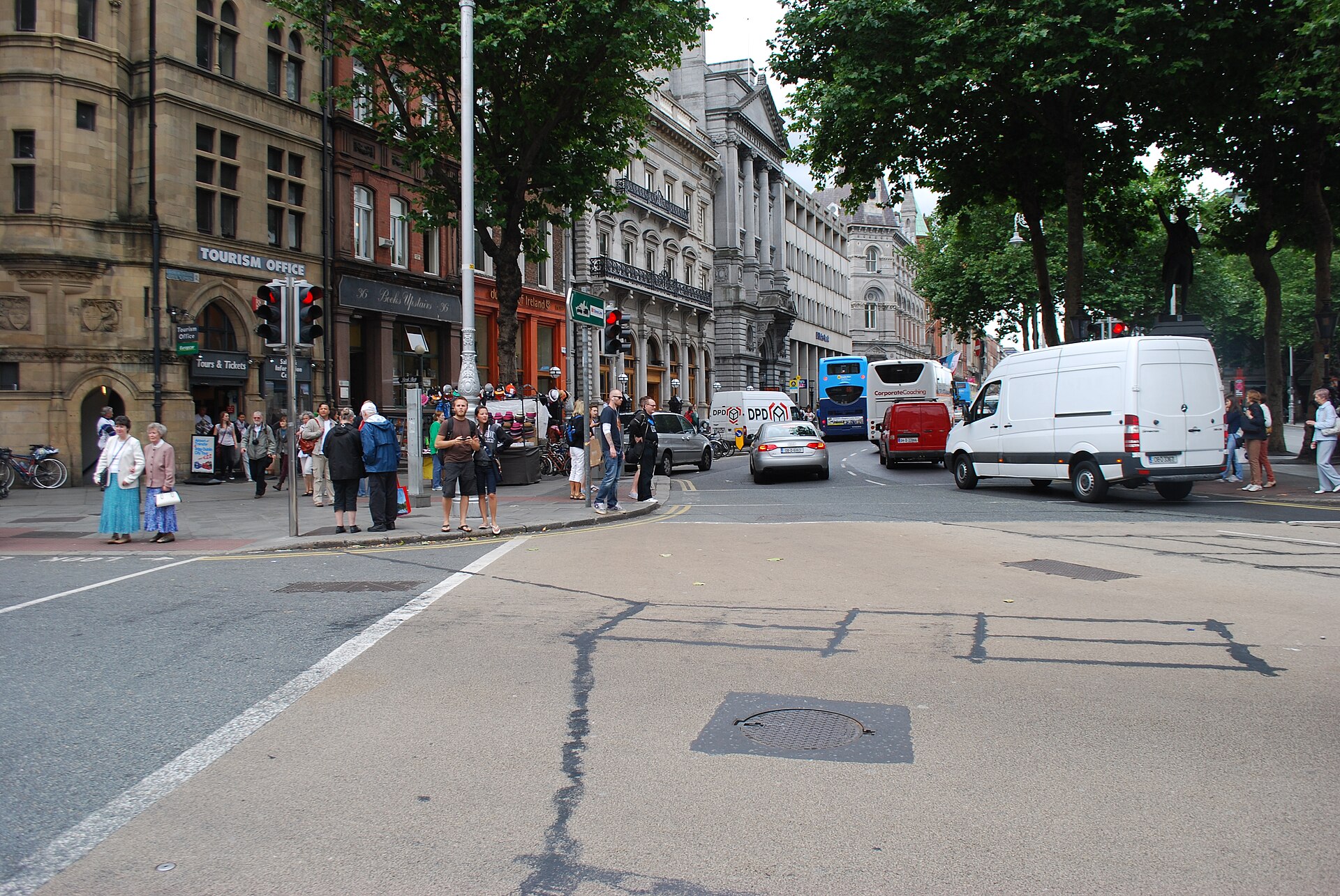Pedestrians wait at a crossing on College Green in Dublin while buses, cars and a white van pass through.