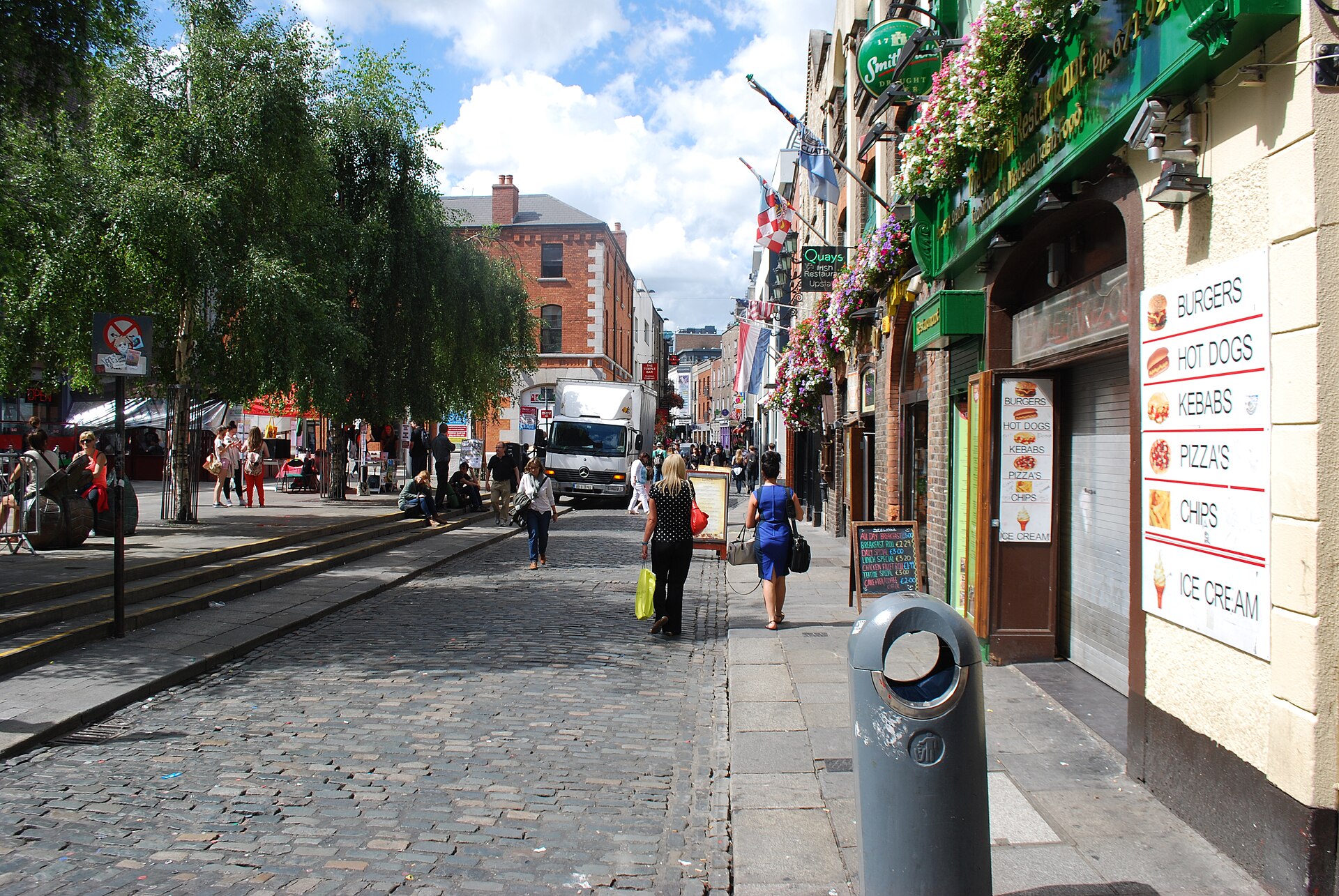 Pedestrians walk along Temple Bar Square in Dublin beside flower-covered pub fronts and a takeaway sign listing food.