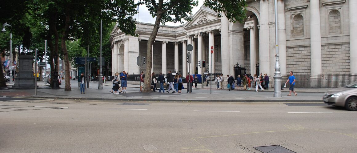 Pedestrians walk beside the columned former Parliament House at College Green in Dublin, shaded by leafy street trees.