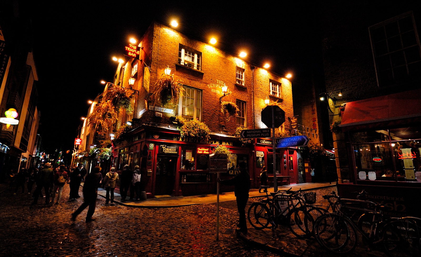 Pedestrians walk past the illuminated Temple Bar pub in Dublin at night, with bicycles beside the cobbled street.