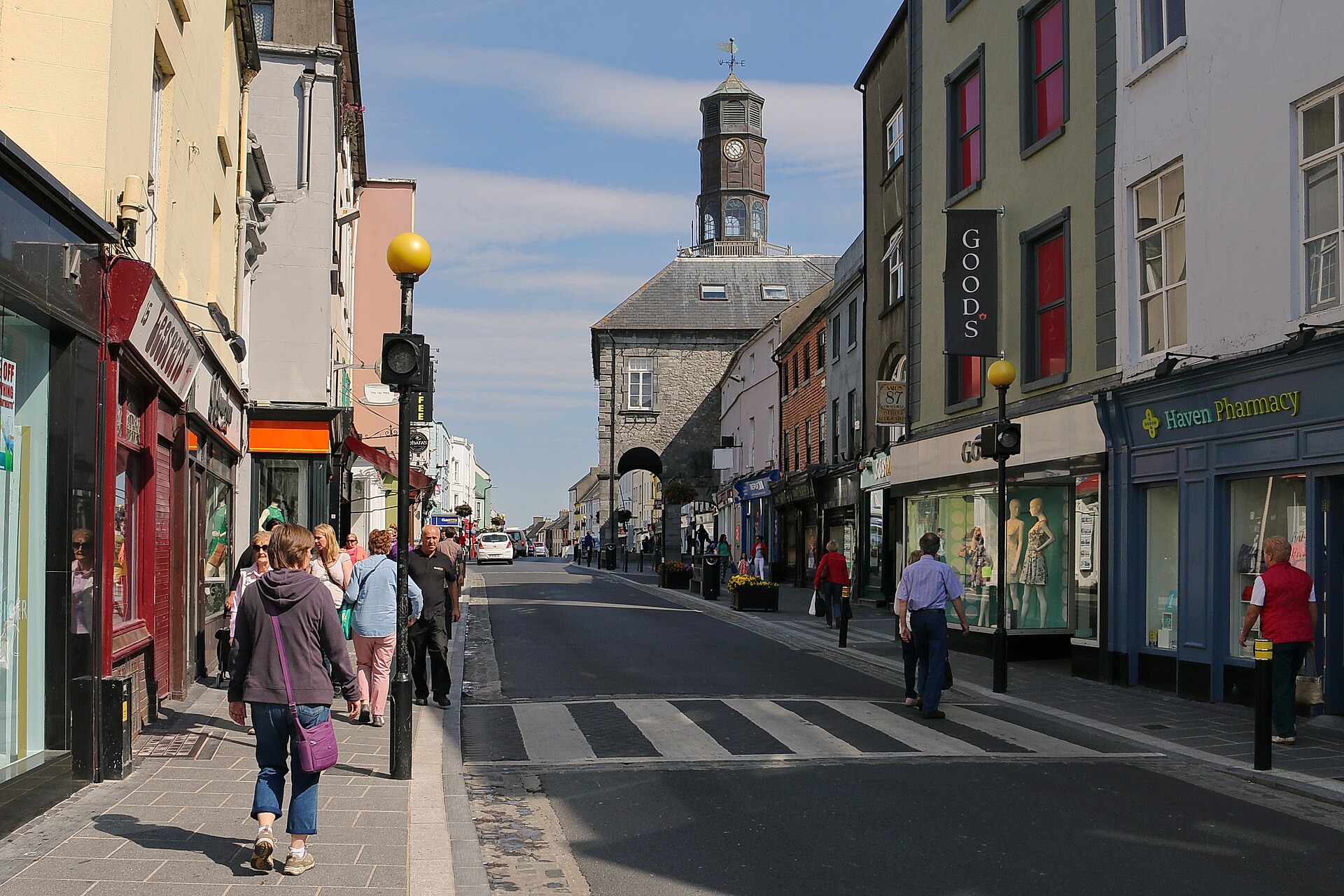 Pedestrians walk beside shops on Kilkenny's High Street, where a zebra crossing spans the road below a distant clock tower.