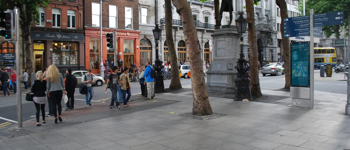 People walk beneath trees beside the Grattan statue at College Green in Dublin, with traffic and shopfronts behind.