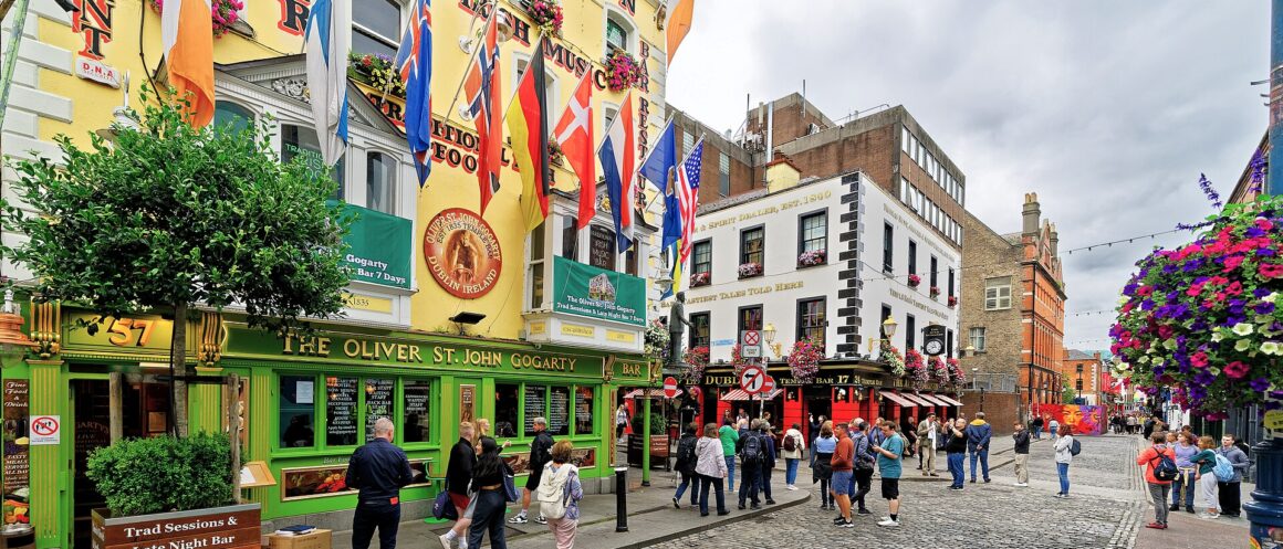 People walk past colourful restaurant and bar fronts, flags and hanging flower baskets on Temple Bar Square in Dublin.