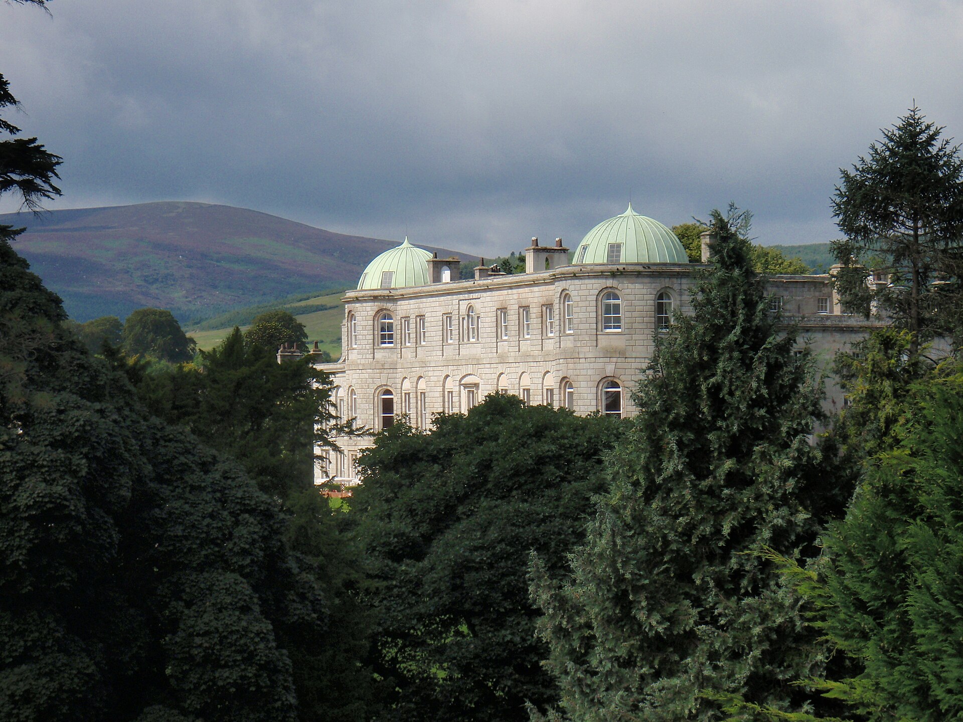 Powerscourt House and its paired green domes rise above garden trees, with a broad mountain slope behind.