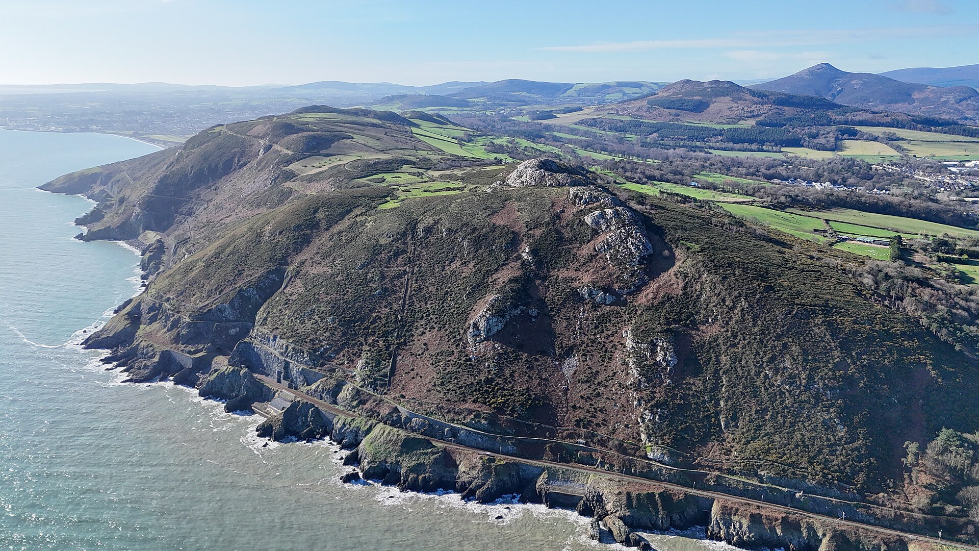 The railway follows rocky cliffs below Bray Head, with green fields and hills stretching inland from the coast.