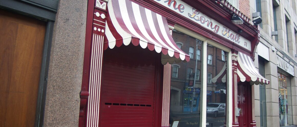 Red frontage of the Long Hall in Dublin, with striped awnings above its window and closed doorway.