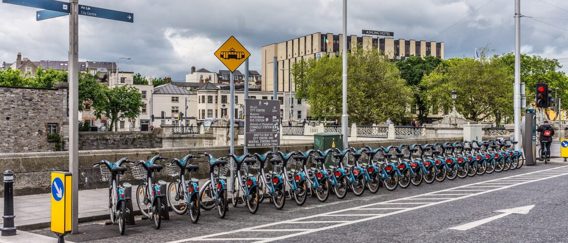 A row of DublinBikes bicycles docked beside the Liffey near Heuston Station, with buildings and tram wires behind.
