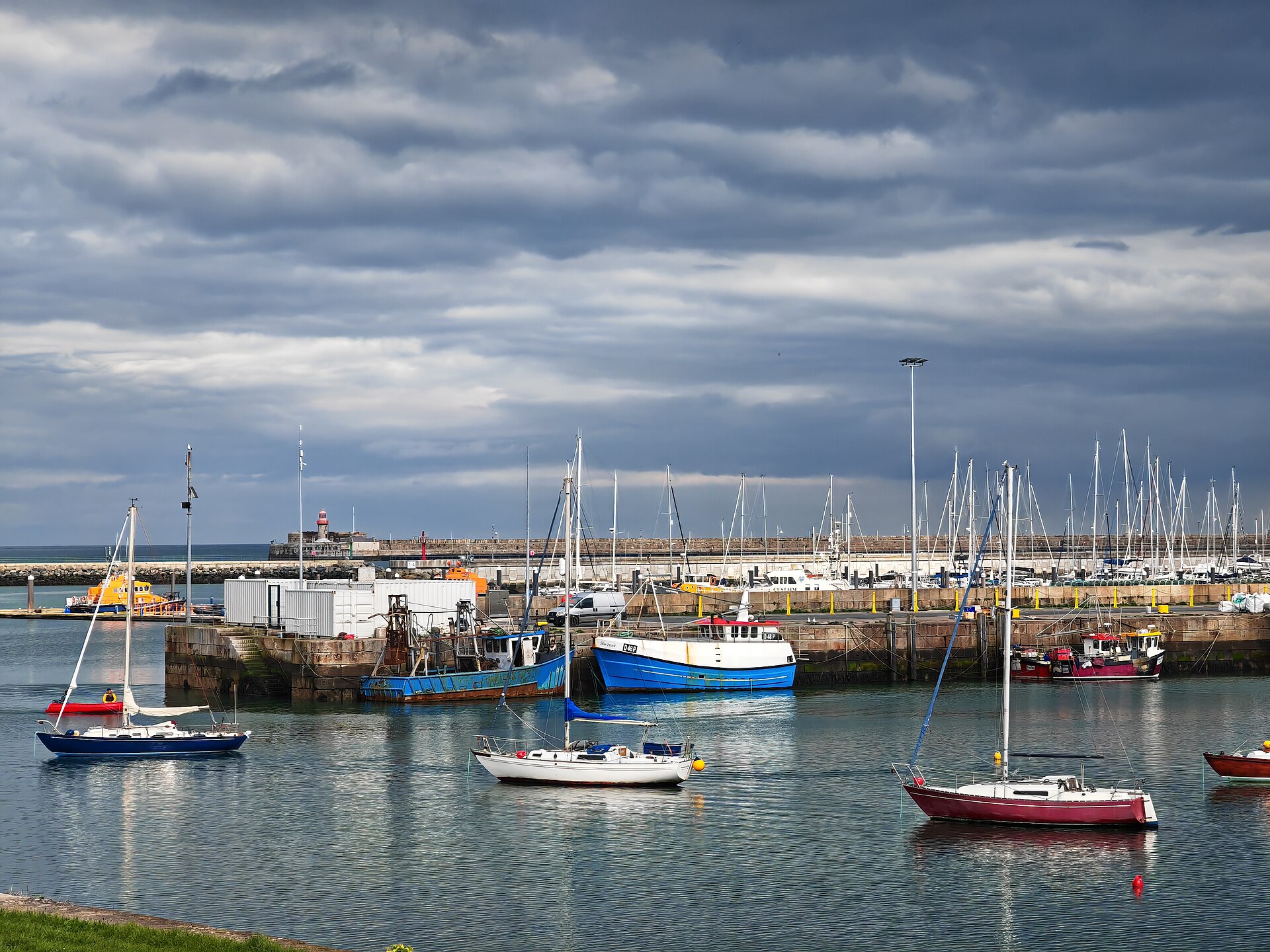 Sailing yachts lie at moorings in Dun Laoghaire Harbour, with fishing boats, marina masts and a lighthouse beyond.