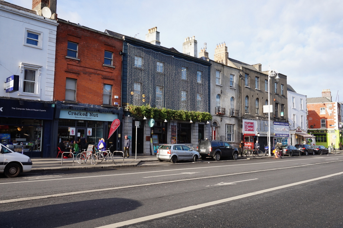 Shopfronts and pubs line Camden Street Lower in Dublin, with bicycles, parked cars and pedestrians along the pavement.