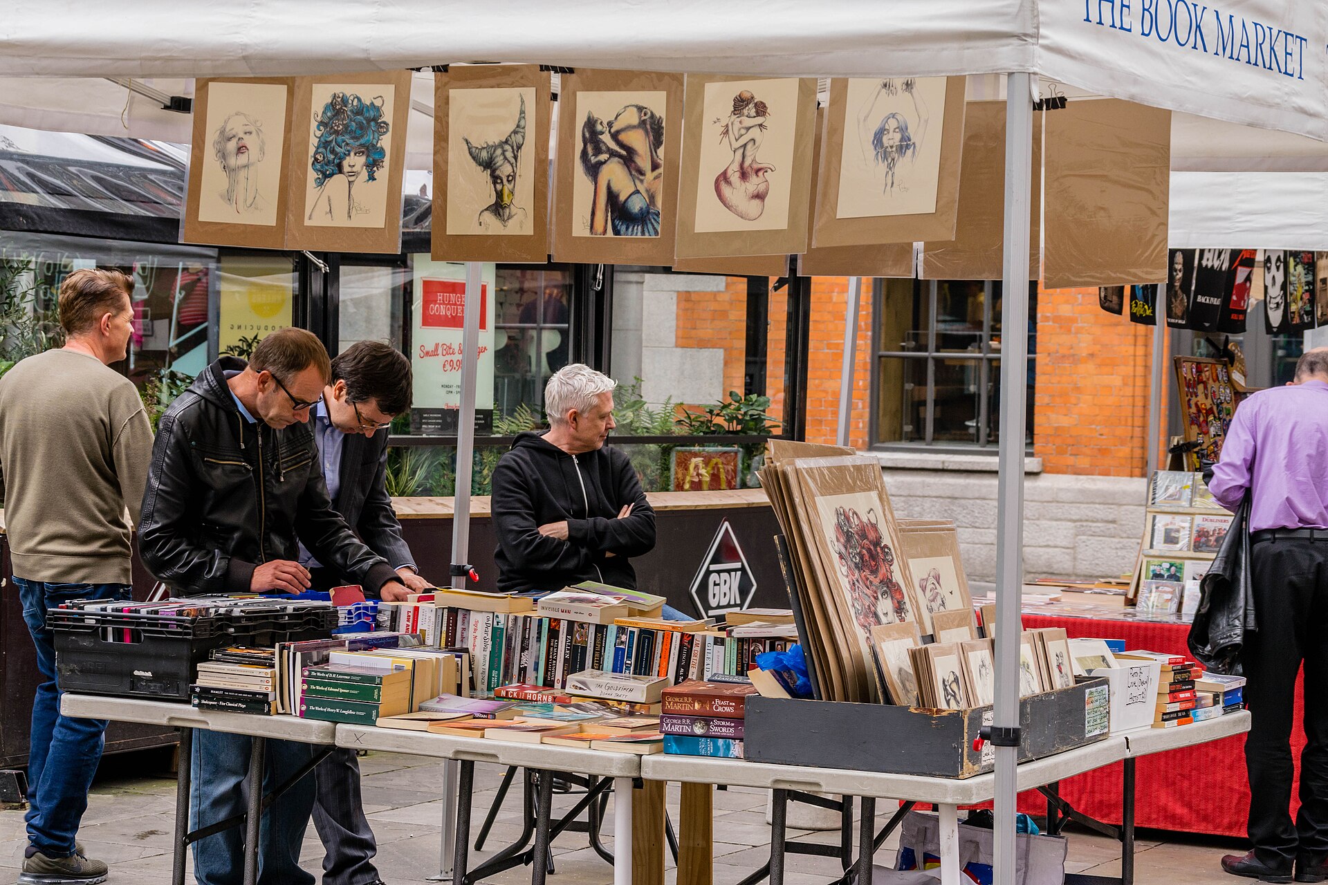 Shoppers browse books and illustrated prints beneath a white canopy at a market stall in Temple Bar Square, Dublin.
