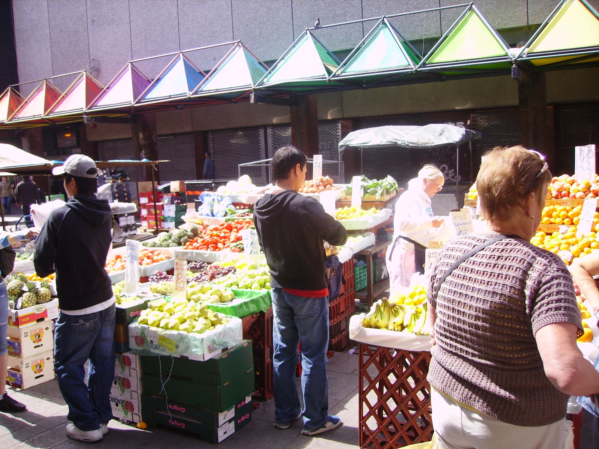 Shoppers browse boxes of fruit and vegetables beneath umbrellas at Moore Street market in Dublin on a sunny day.