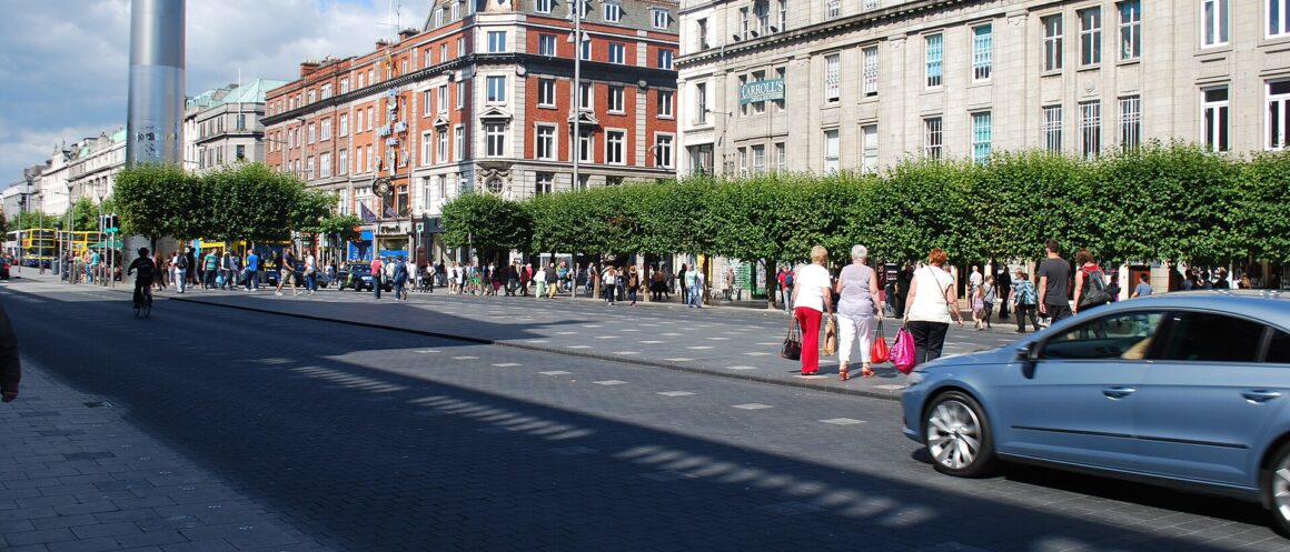 Shoppers cross O'Connell Street in Dublin beneath the Spire, with trees lining the opposite pavement.