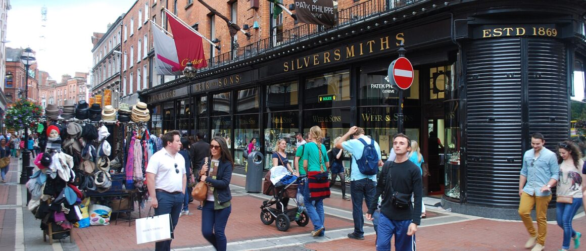 Shoppers pass a hat stall and a black corner shopfront at the junction of Wicklow Street and Grafton Street, Dublin.