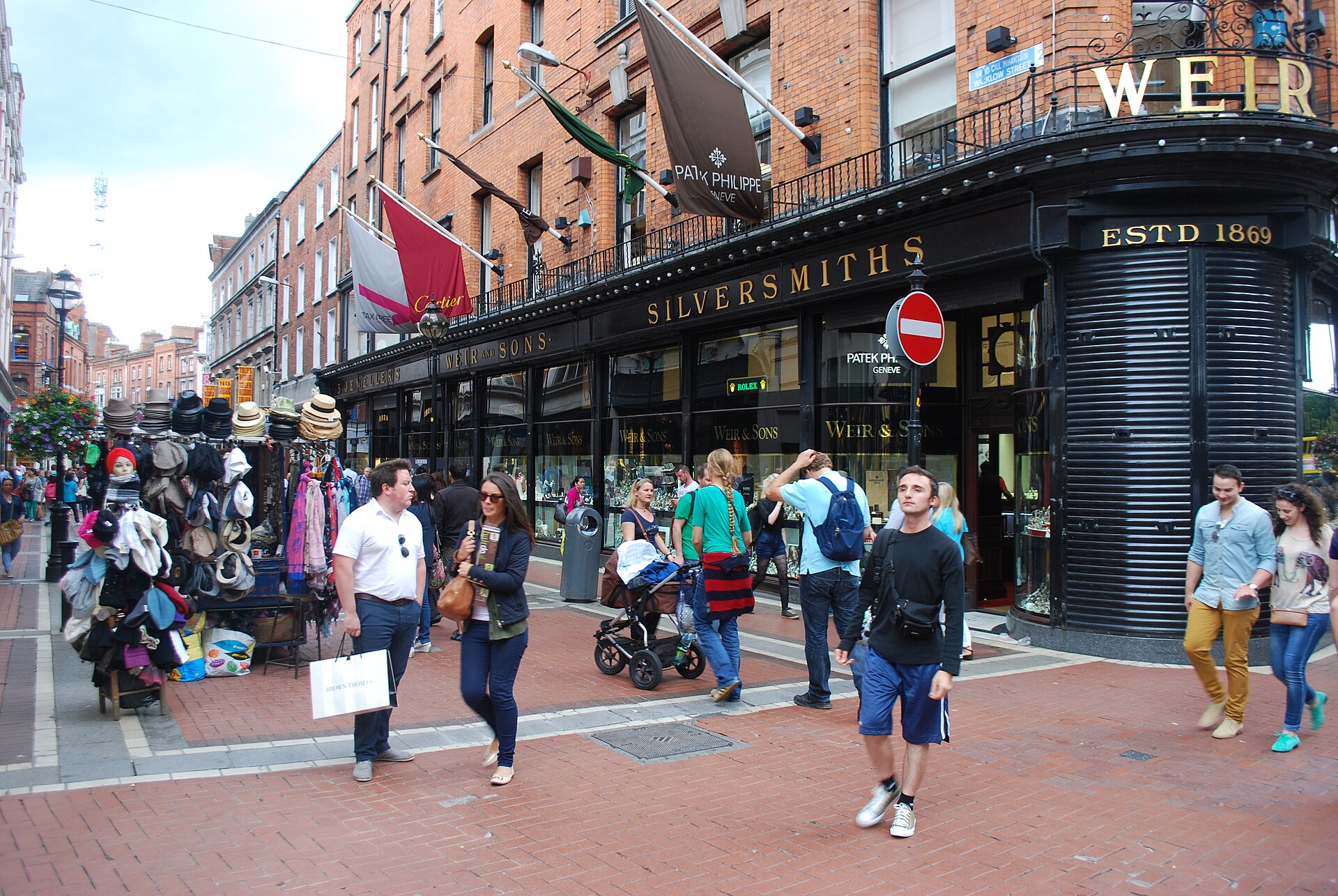 Shoppers pass a hat stall and a black corner shopfront at the junction of Wicklow Street and Grafton Street, Dublin.