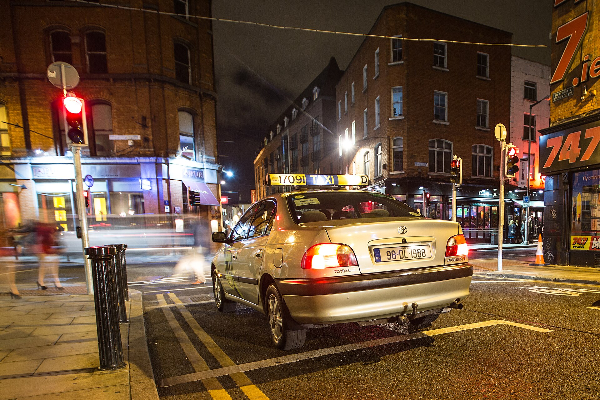 A silver Dublin taxi waits at a red light on a city street illuminated by shopfronts at night.
