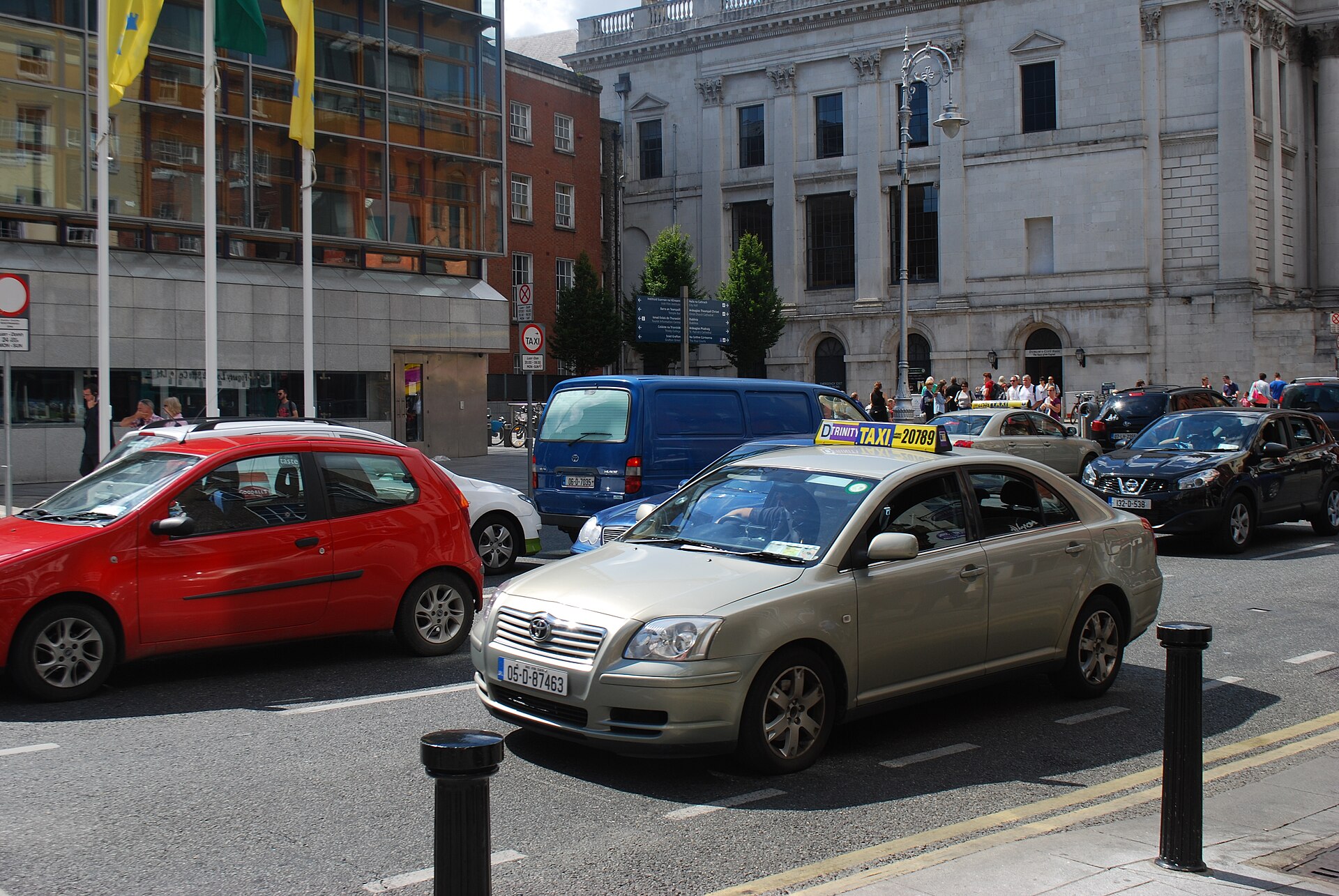 A silver Dublin taxi with a yellow and blue roof sign passes City Hall on busy Dame Street.