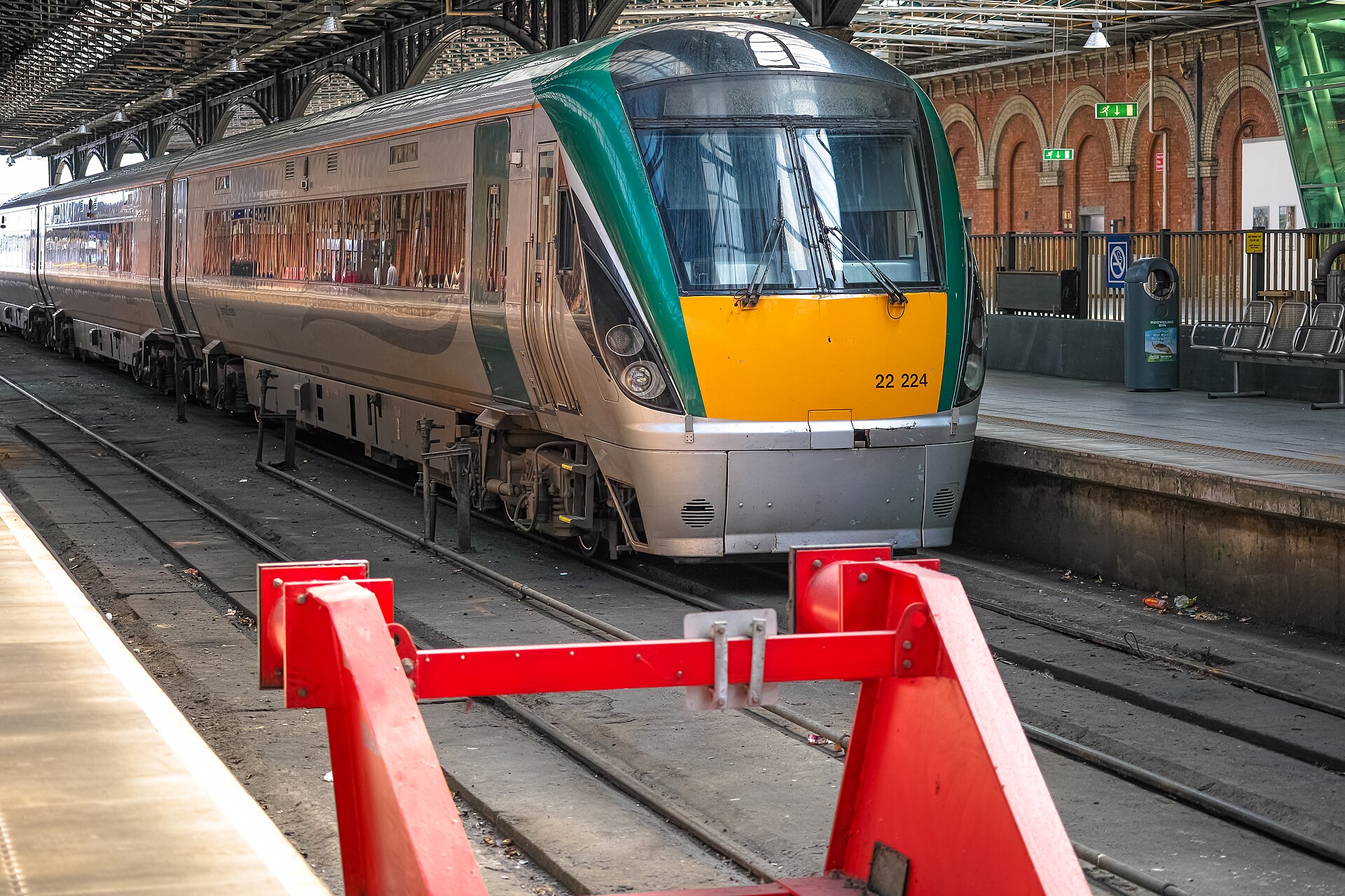 A silver and green Irish Rail InterCity railcar stands behind red buffers beneath Dublin Connolly station's roof.
