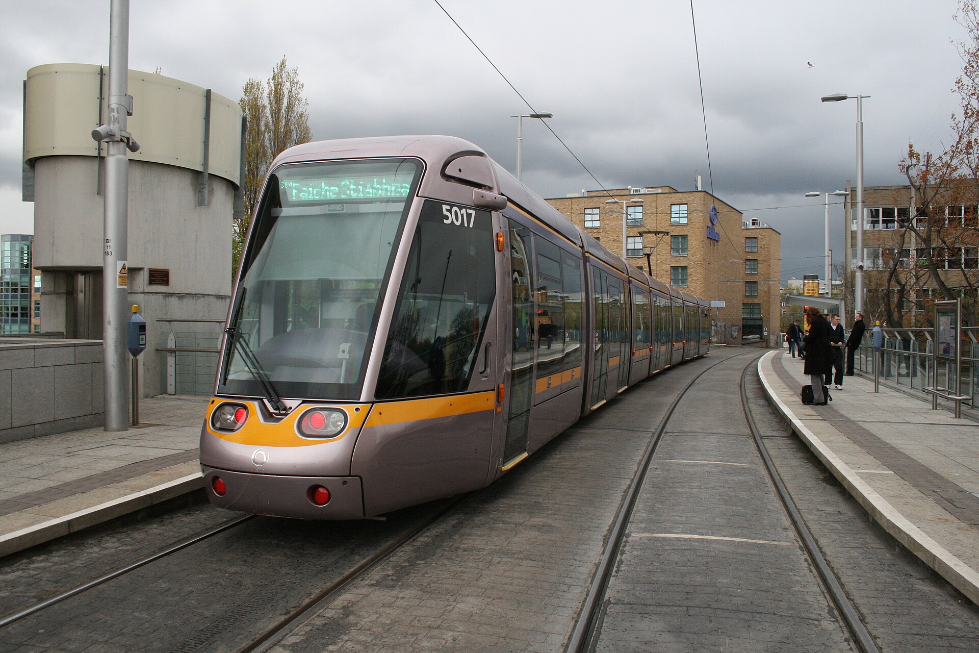 A silver and purple Luas tram beside the curved platform at Charlemont in Dublin, with passengers waiting.