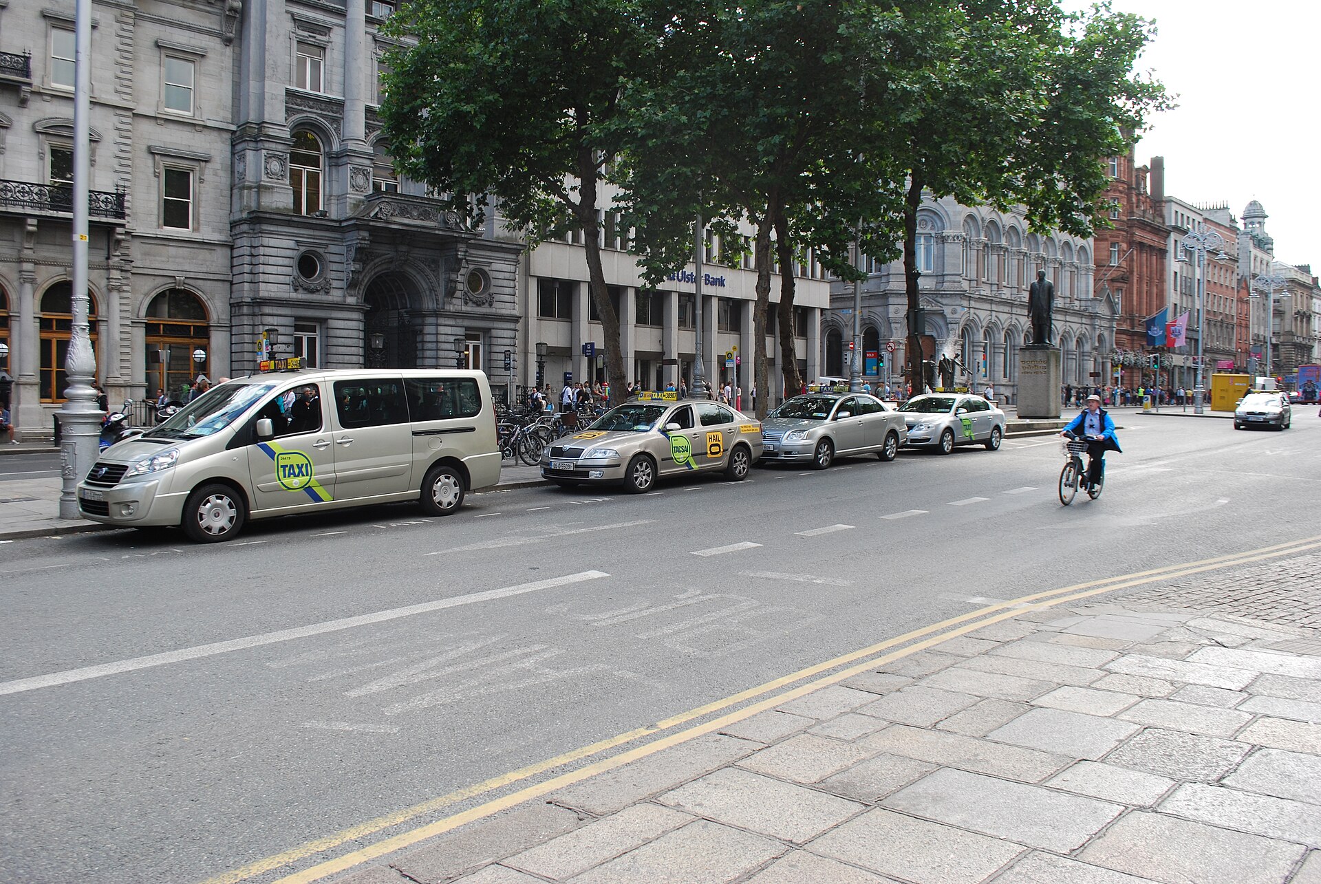 A silver taxi van and several sedan taxis line the kerb on Dame Street in central Dublin.