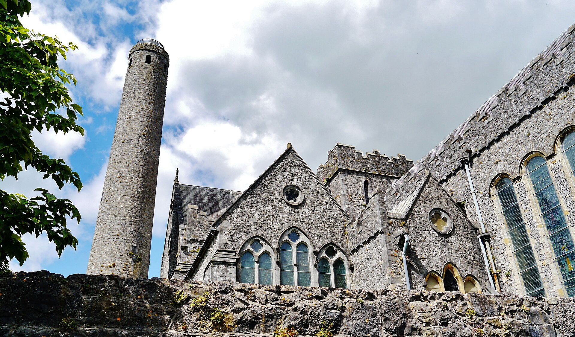 St Canice's Cathedral's pointed windows and stone gables stand beside its tall round tower in Kilkenny beneath a cloudy sky.