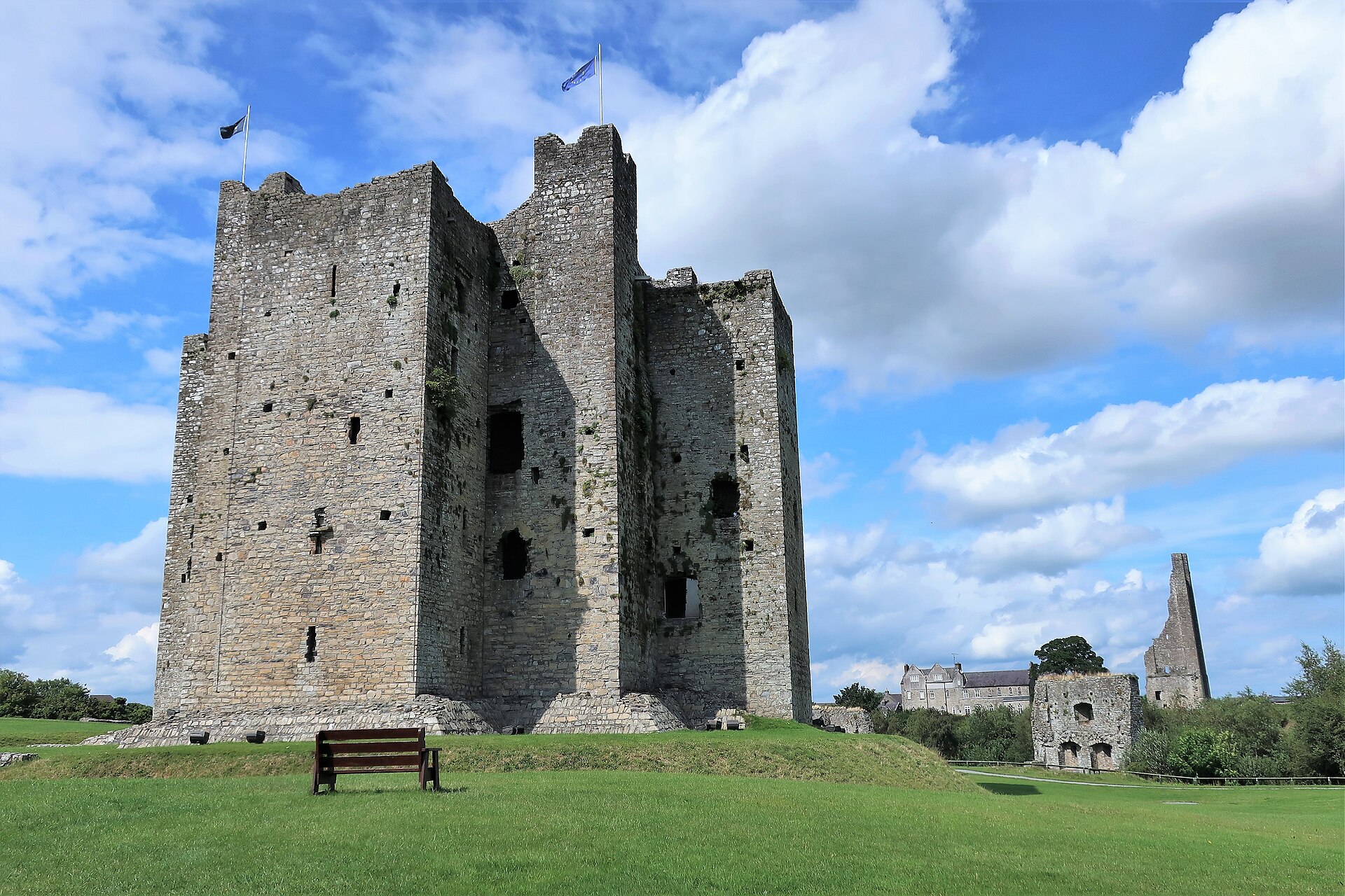 The stone keep of Trim Castle towers above a green lawn and wooden bench, with flags flying against blue sky.