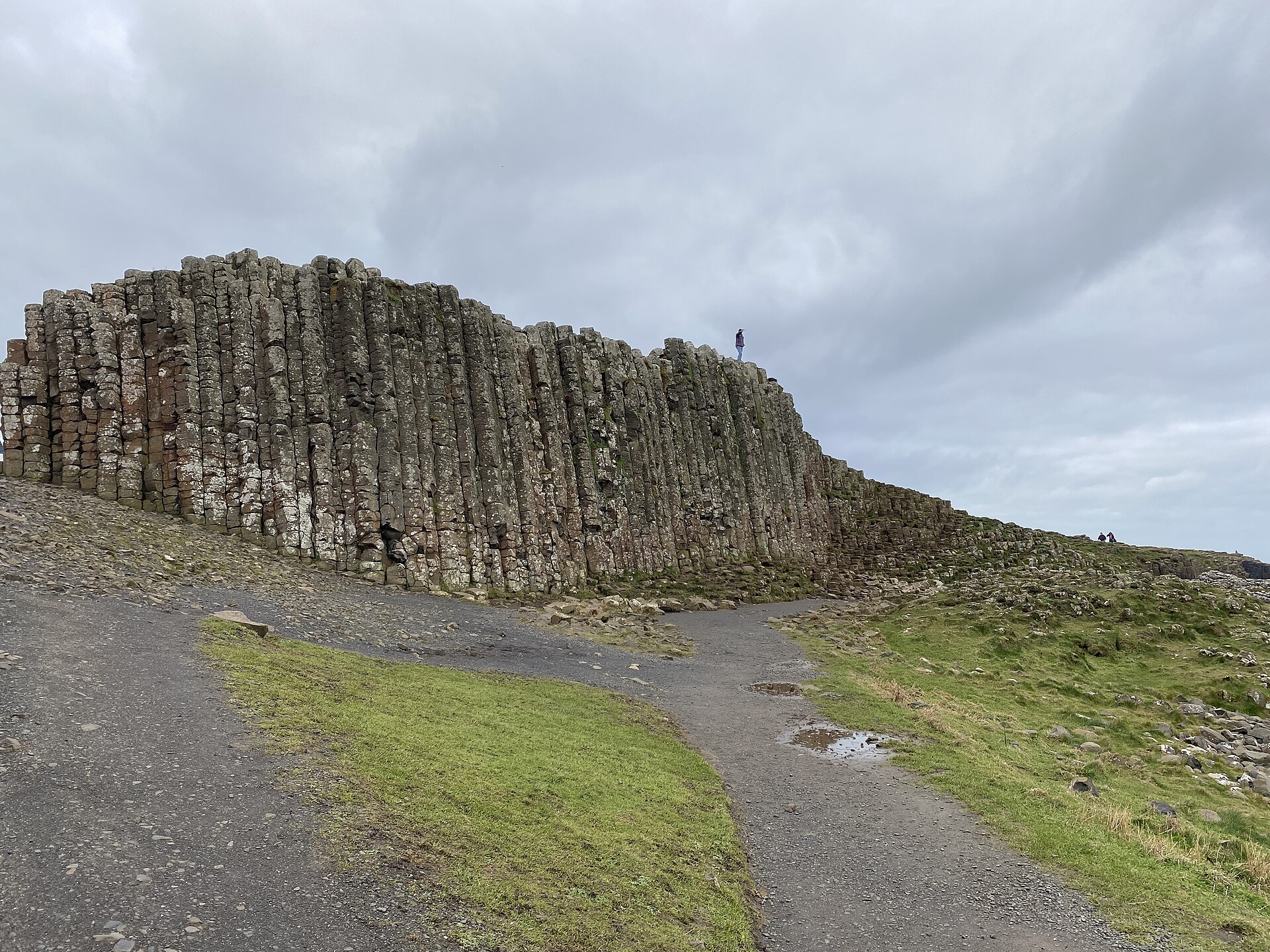 Tall basalt columns form a dark stepped wall at the Giant's Causeway, above a gravel path and patches of grass.