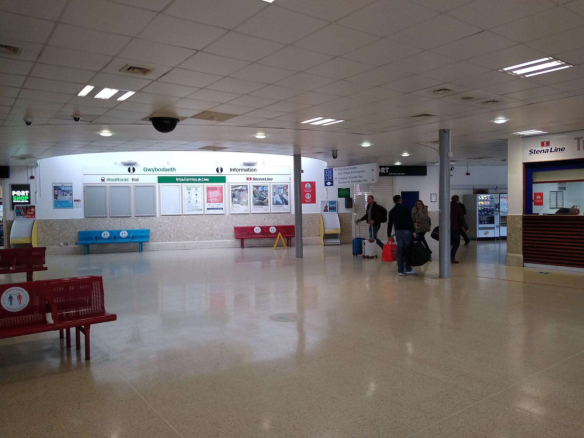 Travellers with luggage cross the concourse of the combined railway station and ferry terminal at Holyhead, beneath Rail, Irish Ferries and Stena Line information boards