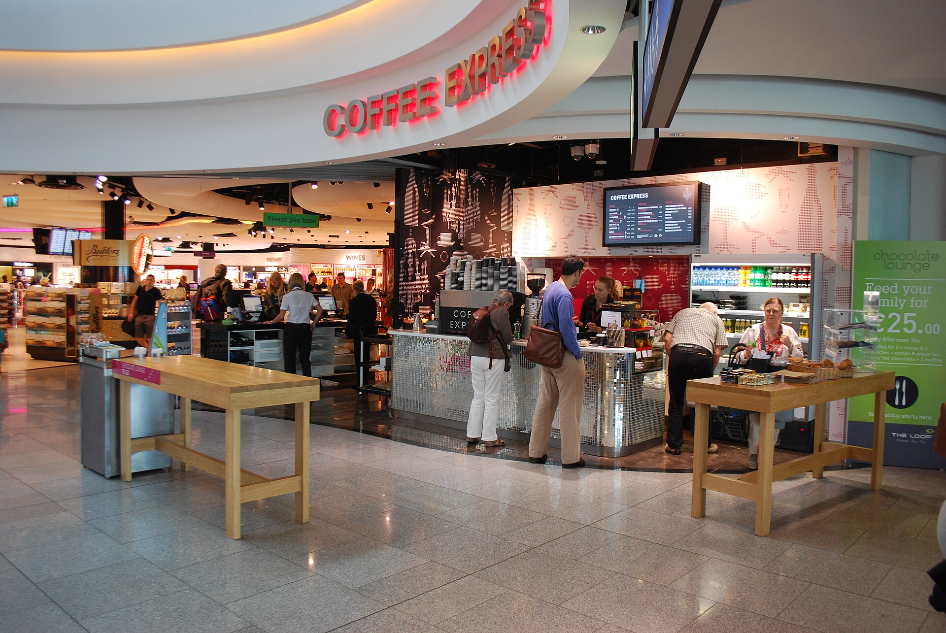 Travellers sit and stand at tall cafe tables beside a coffee counter in Dublin Airport Terminal 2.