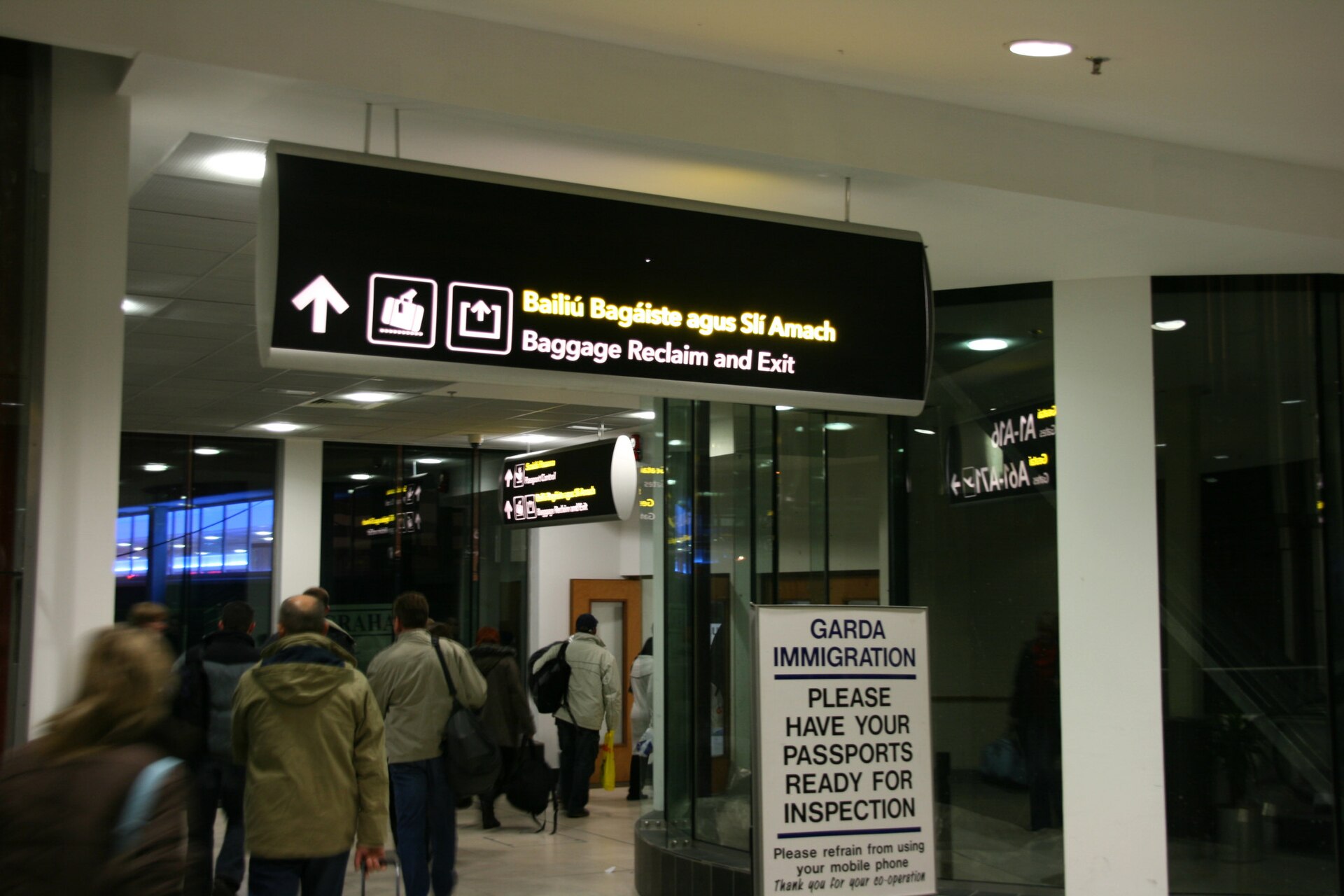Travellers walk through Dublin Airport arrivals beneath baggage-reclaim signs, beside a notice asking for passports ready for inspection.