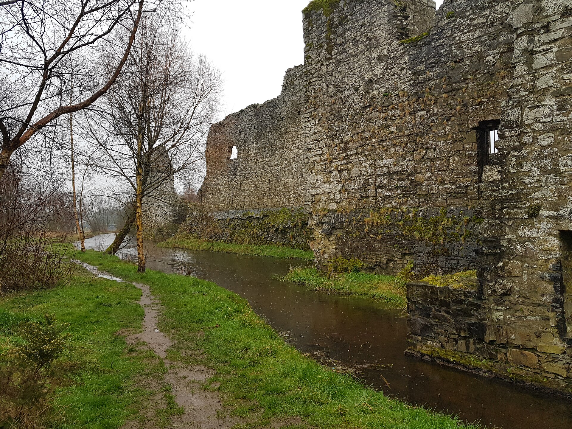 Trim Castle curtain walls rise beside water and a narrow grassy path, with bare trees along the outer edge.