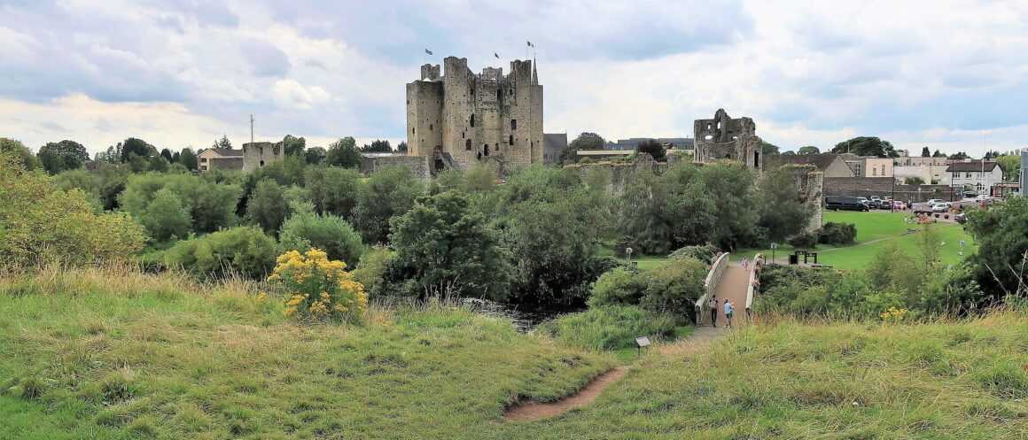 Trim Castle rises beyond the River Boyne and a footbridge, with grassy riverbanks and visitors in the foreground.