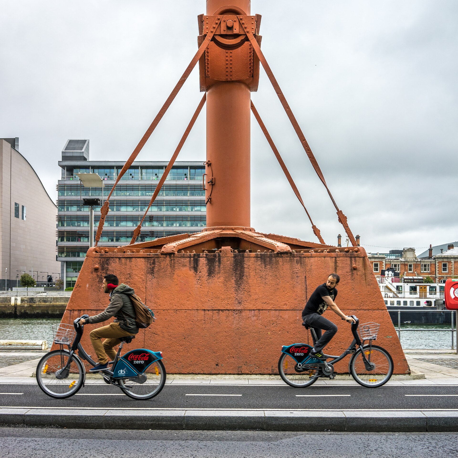 Two DublinBikes cyclists pass the orange diving bell beside the River Liffey on Dublin's quays under a cloudy sky.