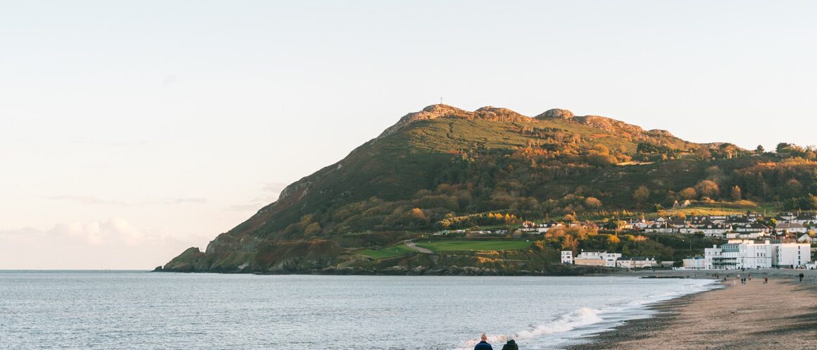 Two walkers follow Bray beach beneath the sunlit slopes of Bray Head, with seafront buildings beside the water.