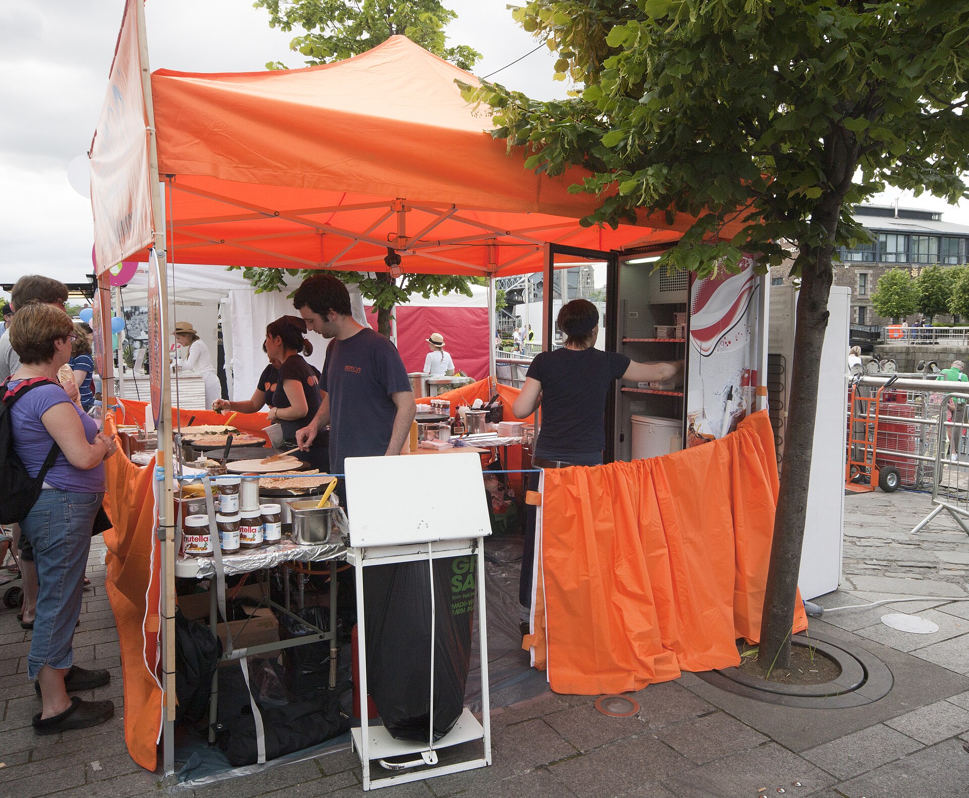 Vendors prepare food beneath an orange canopy at the Dublin Maritime Festival market, with jars and utensils along the counter.