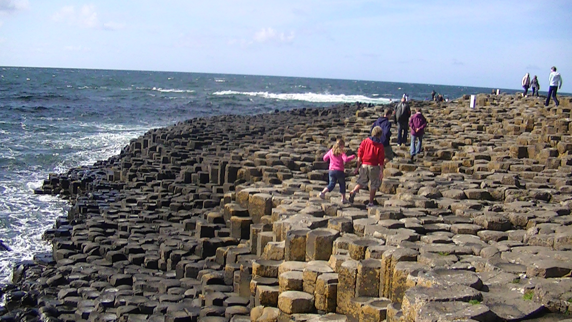 Visitors cross the stepped basalt columns at the Giant's Causeway, with waves breaking against the dark shoreline beside them.