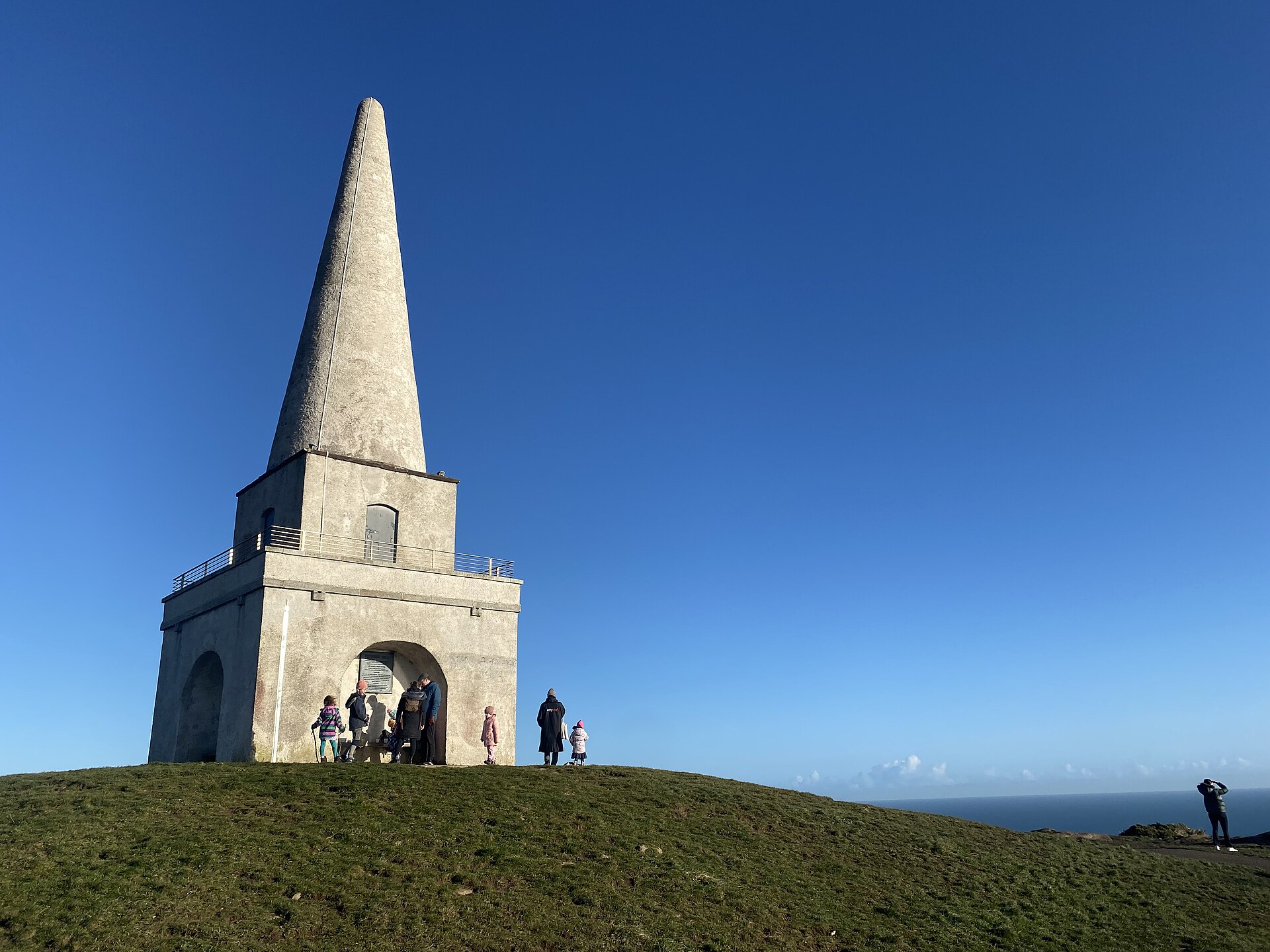 Visitors gather around the arched base of the Killiney Hill obelisk on a grassy summit above the sea.