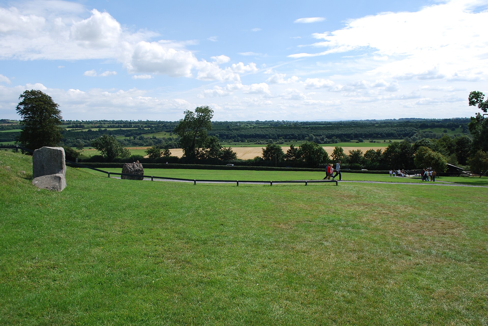 Visitors walk along a path at Newgrange above the green fields and wooded countryside of the Boyne Valley.