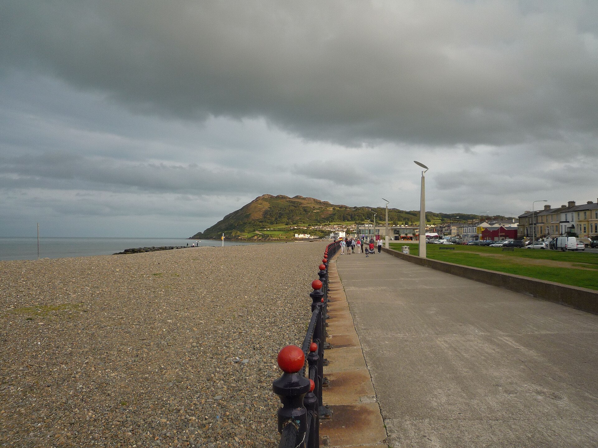 Walkers follow Bray seafront promenade beside the shingle beach, with Bray Head ahead beneath a cloudy sky.