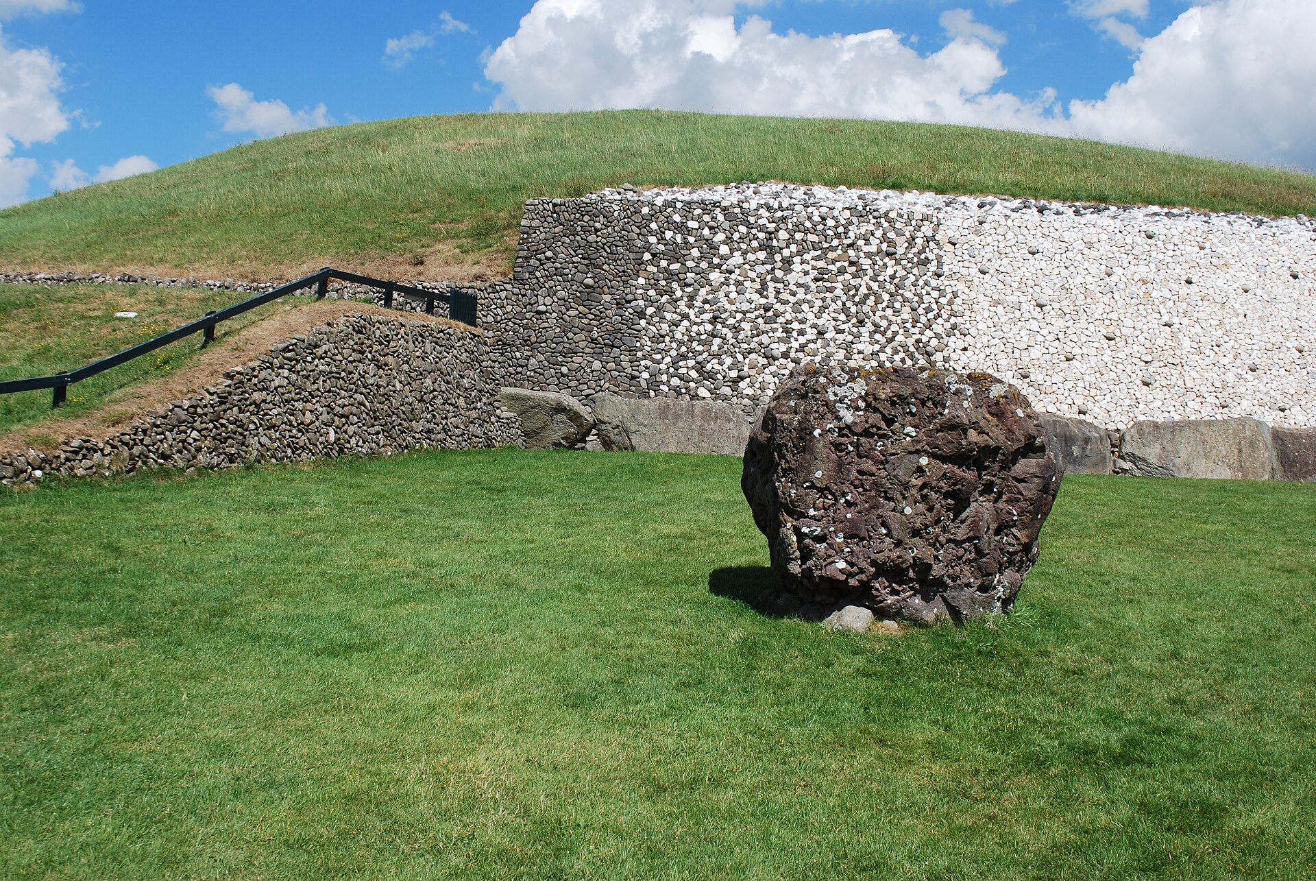 A weathered standing stone sits on the lawn beside Newgrange, with the white quartz facade and grassy mound behind.