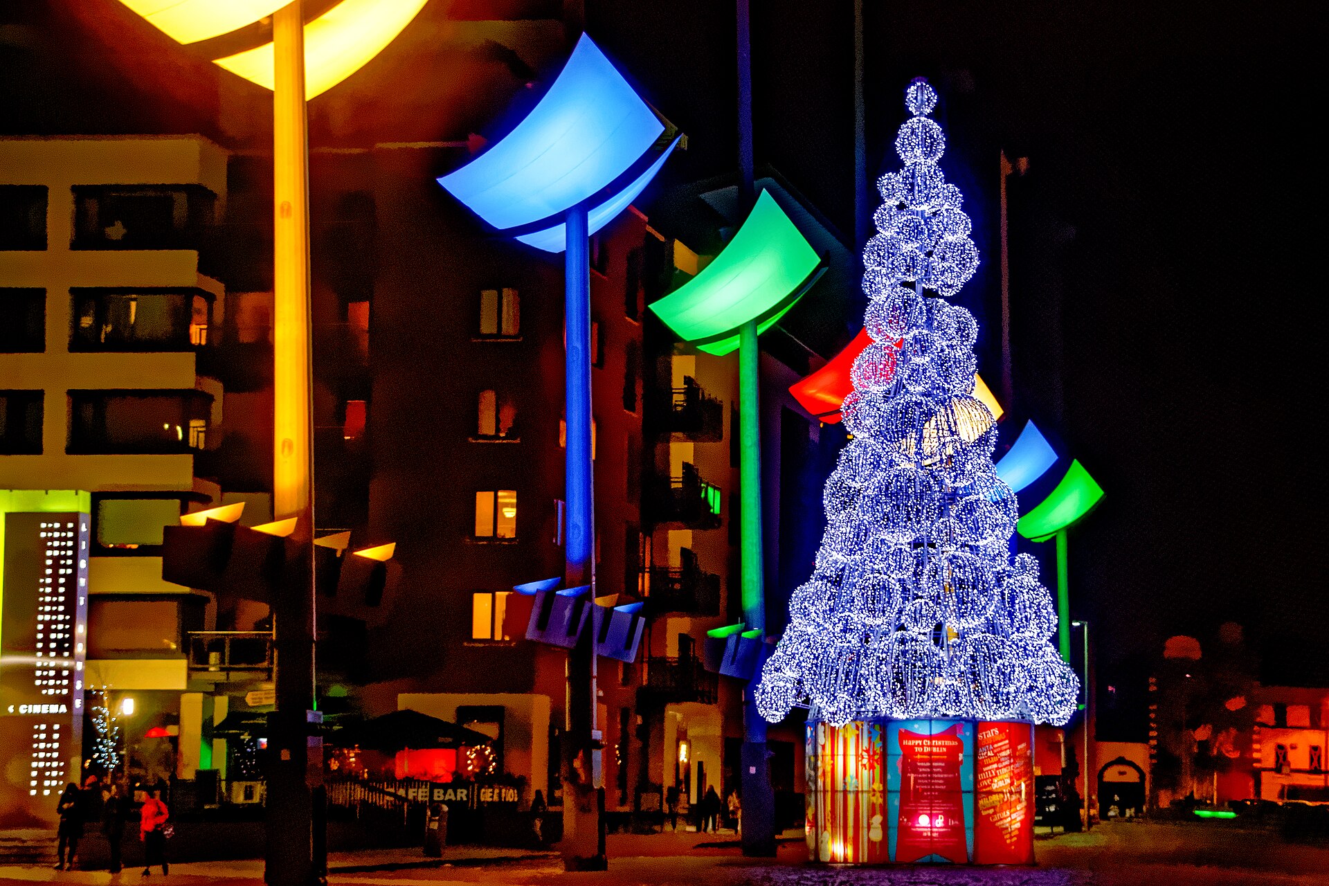 A white Christmas tree glows beside colourful street lights in Dublin’s Smithfield square, with apartments behind it at night.