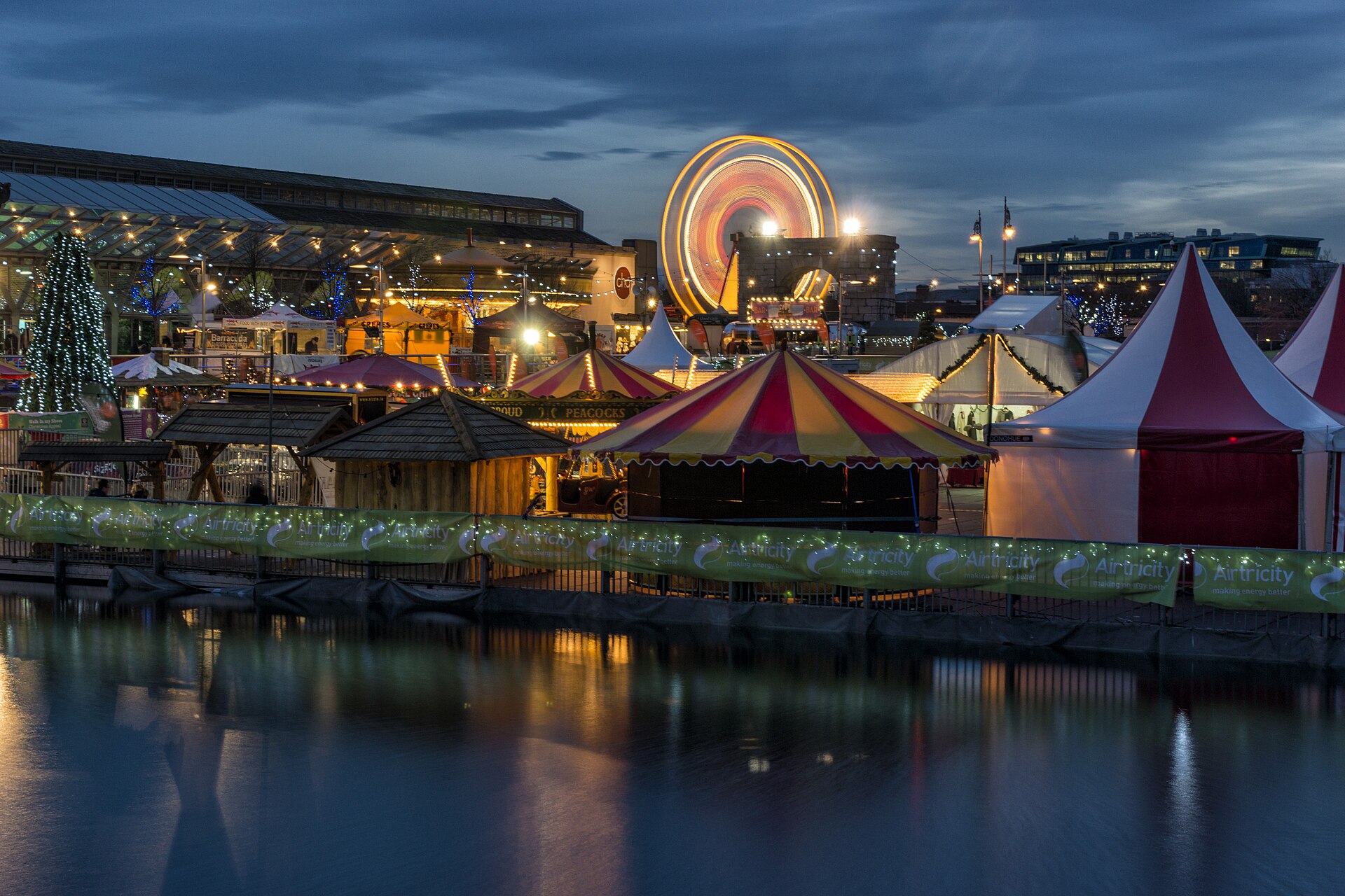 Wooden stalls and striped tents line the water beside a glowing Ferris wheel at Dublin's Docklands Christmas Festival.