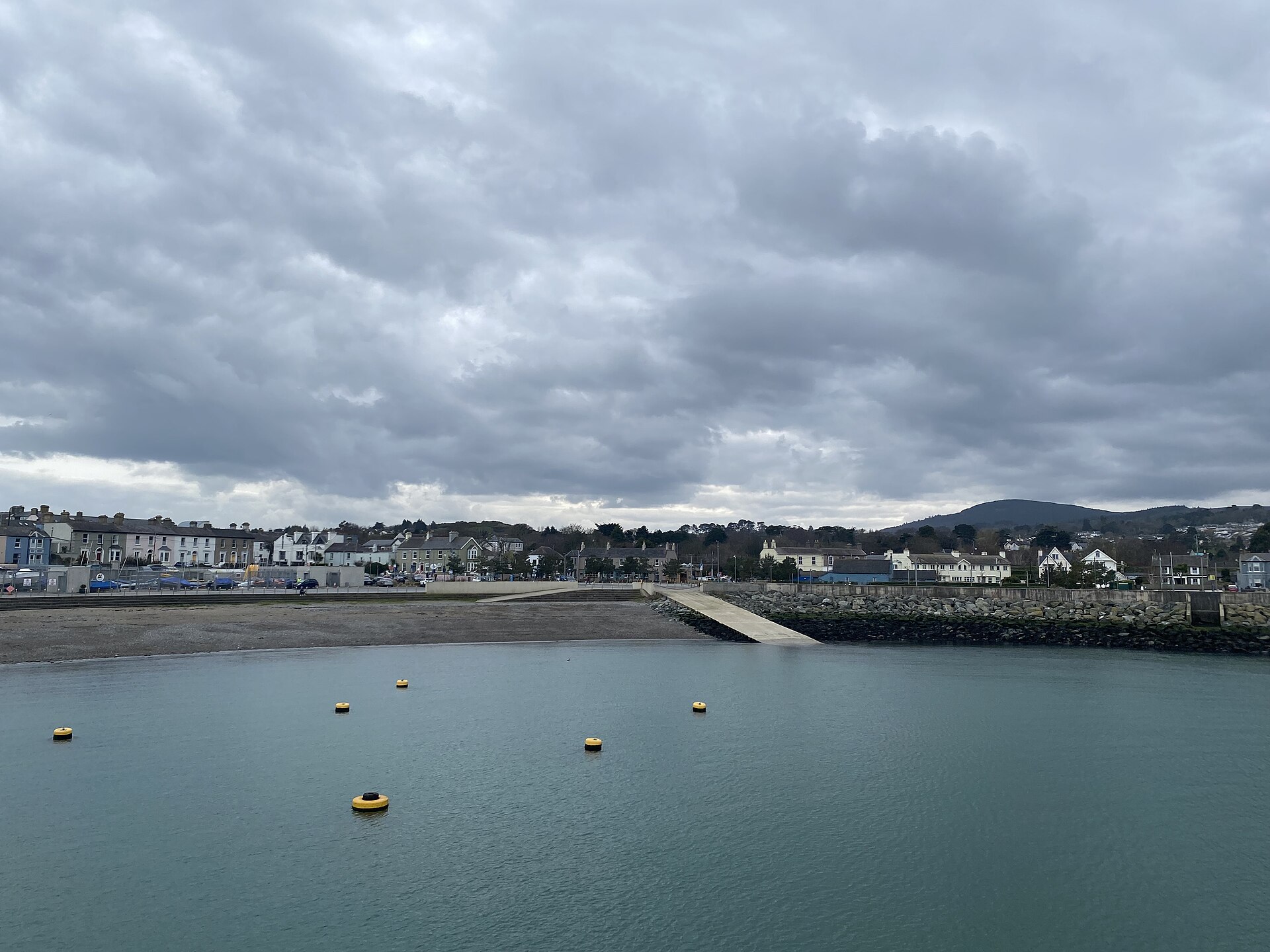 Yellow buoys float in Greystones Harbour below a cloudy sky, with a beach, slipway and waterfront buildings across the water.